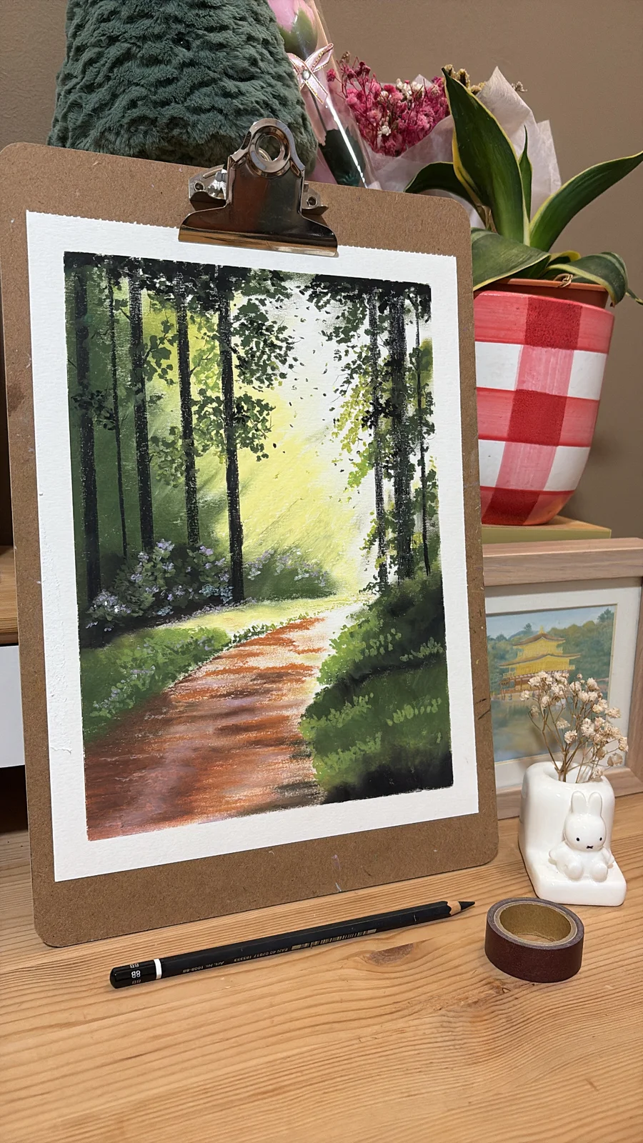 Painting of a sunlit path winding through a green forest, displayed on a wooden easel.
